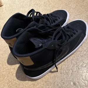 Svarta Nike sneakers - Snygga svarta Nike sneakers med högt skaft och vit sula. Skorna har snörning och en stilren design med en diskret Nike-logga på sidan. Perfekta för en casual look.