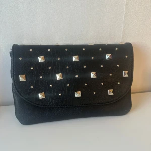 Svart axelväska med nitar - Snygg svart axelväska i skinn med silverfärgade nitar på framsidan. Väskan har en avtagbar axelrem och en stilren design som passar perfekt för en trendig look.