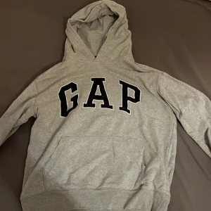 Grå hoodie från GAP - Säljer en klassisk grå hoodie från GAP med stor logga på bröstet. Tröjan har en bekväm passform med känguruficka och ribbade muddar. Perfekt för en avslappnad stil.
