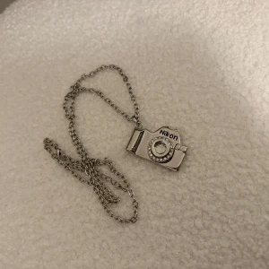 Kamerahalsband med Nikon-design - Unikt halsband med en kamerahänge i silverfärg, prytt med gnistrande stenar runt linsen. Perfekt för fotointresserade som vill bära sin passion med stil. Kedjan är tunn och elegant, vilket gör det lätt att bära till olika tillfällen.