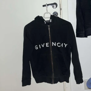 Svart hoodie från Givenchy - Säljer en stilren svart hoodie från Givenchy med vit logotyp på framsidan och ett grafiskt mönster på baksidan. Tröjan har dragkedja och långa ärmar, perfekt för en casual look. 100% äkta direkt köpt från NK barnavdelning Nypris 2500kr