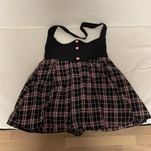 Kawaii rutig kjol med hjärtknappar - Kjol med rutigt, rosa och svart kjolparti. Klänningen har söta hjärtformade knappar framtill. Storlek L, Mått: byst-90-104cm, midja-82cm, längd: 91/60cm.