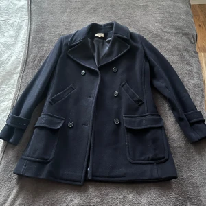 Mörkblå kappa med knappar - Elegant mörkblå kappa med dubbelknäppning och stora fickor framtill. Klassisk design med långa ärmar och bred krage. Perfekt för kyligare dagar.