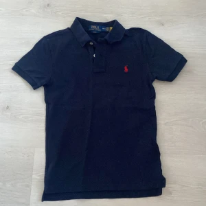 Mörkblå pikétröja från Ralph Lauren - Säljer en stilren mörkblå pikétröja från Ralph Lauren med klassisk krage och knappar. Tröjan har en röd broderad logotyp på bröstet och är i slim fit. Perfekt för en avslappnad men ändå uppklädd look.