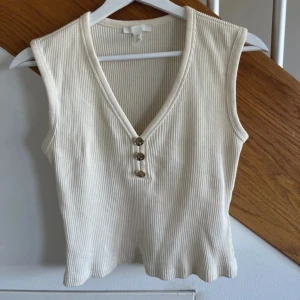 Vit ribbad linnetopp från H&M - Säljer en ljusbeige ribbad linnetopp från H&M. Toppen har en v-ringning med tre guldiga knappar framtill. 