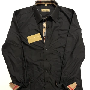 Svart skjorta från burburry  - Snygg svart skjorta från Burrbury med klassisk krage och knappar framtill. Perfekt för en stilren look. Skjortan har långa ärmar och en diskret broderad logga på bröstet. Du köper den med  en liten defekt på bild 5 om inte ens syns.