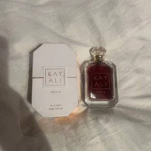 Kayali Vanilla 28 Eau de Parfum - Elegant parfym från Kayali med en rik och djup vaniljdoft. Flaskan är stilrent designad i glas med en facetterad kork och en mörk bärnstensfärgad vätska. Förpackningen är vit med guldiga detaljer, vilket ger en lyxig känsla.