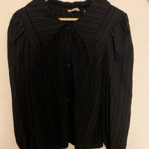 Svart randig blus - Snygg svart blus med diskreta ränder och knappar framtill. Blusen har långa ärmar och en klassisk krage som ger en stilren look. Perfekt för en elegant touch till din outfit.