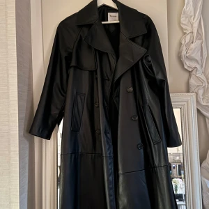 Svart trenchcoat i skinnimitation - Snygg svart trenchcoat i skin imitation  från Bershka. Jackan har en klassisk design med knappar och långa ärmar. Perfekt för en stilren look. Ett skärp kommer till också :)
