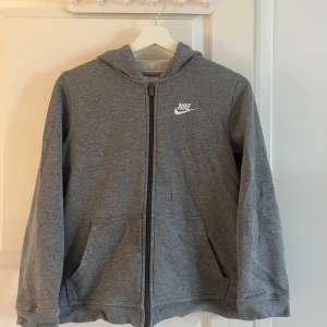 Grå hoodie från Nike - Säljer en grå hoodie från Nike med dragkedja och huva. Den har en klassisk design med Nike-logga på bröstet och praktiska fickor framtill. Perfekt för en avslappnad stil.