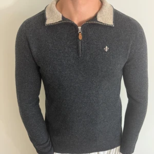Morris Quarter Zip S/M - Säljer en stilren grå stickad tröja från Morris med en broderad detalj på bröstet. Tröjan har en dragkedja vid halsen och en kontrasterande beige krage. Perfekt för kyligare dagar! Använd fåtal gången. Obs står xl men passar s/m