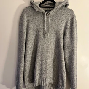 Grå stickad hoodie Dressman - Säljer en mysig grå stickad hoodie med huva och dragsko. Perfekt för alla väder. Den har långa ärmar och ribbstickade muddar vid ärmslut och nederkant.