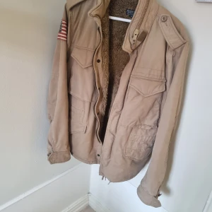 Ralph lauren field jacka - Säljer en beige jacka från Ralph Lauren med teddyfoder och amerikansk flagga på ärmen. Jackan har dragkedja och knappar framtill samt flera fickor. Perfekt för kyliga dagar!