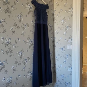 Marinblå balklänning - Elegant marinblå långklänning från Lipsy med glittrig överdel och offshoulder-design. Klänningen har en slits som ger en extra touch av stil. Perfekt för speciella tillfällen.