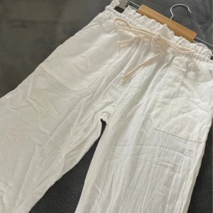 Vita linnebyxor med snörning - Snygga vita linnebyxor med elastisk midja och snörning för en avslappnad look. Byxorna har praktiska fickor fram och är perfekta för varma dagar.