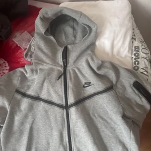 Grå Nike Tech Fleece - Säljer en grå hoodie från Nike med dragkedja och huva. Jackan har en svart Nike-logga på bröstet och svarta detaljer längs dragkedjan och ärmarna. Svaga tecken på användning