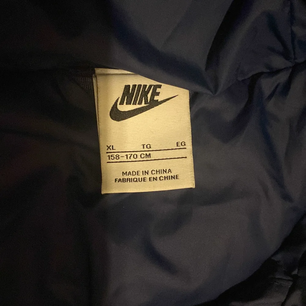 Säljer en snygg mörkblå dunjacka från Nike med huva och dragkedja. Jackan har en klassisk pufferdesign och är perfekt för kyligare dagar. Den har en broderad Nike-logga på bröstet och är både stilren och funktionell.. Takit.