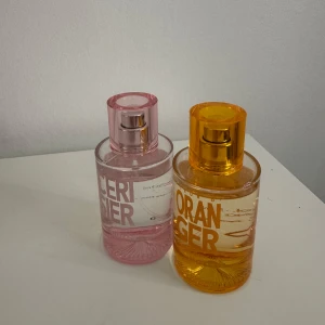 Parfymflaskor i rosa och orange - Två parfymflaskor med färgglada design. Den ena är rosa med texten 'CERISIER' och den andra är orange med texten 'ORANGER'. Båda flaskorna har en rund form och genomskinligt glas som visar vätskan inuti. Perfekt för den som gillar fruktiga och blommiga dofter.