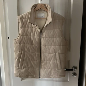 Beige quiltad väst från Bruun & Stengade - Säljer en stilren beige quiltad väst från Bruun & Stengade. Västen har en dragkedja framtill och två praktiska fickor. Perfekt för lager-på-lager nu under våren och kyliga sommarkvällar. Passar både till vardags och mer uppklädda tillfällen. Nypris på denna ligger just nu på runt 1800kr mitt pris 799kr! Modellen är 180cm och storlek M men passar om man har L på vanliga kläder!