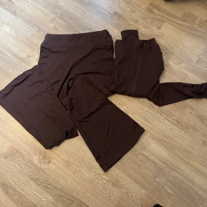 Brun träningsset med dragkedja - Säljer ett brunt träningsset med dragkedja. Setet består av en långärmad topp och matchande byxor. Perfekt för träning eller en avslappnad dag.