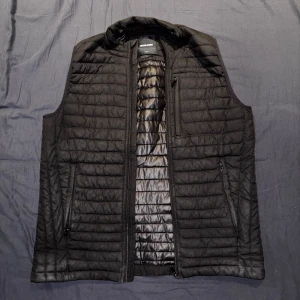 Svart dunväst från Jack & Jones - Säljer en stilren svart dunväst från Jack & Jones. Västen har en quiltad design med dragkedja framtill och praktiska fickor med dragkedjor. Perfekt för lager-på-lager under kyligare dagar.