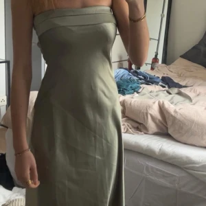 Olivgrön axelbandslös klänning - Snygg olivgrön axelbandslös klänning med en elegant passform. Perfekt för sommarens fester eller en kväll ute. Klänningen har en stilren design med en slät yta och en subtil glans.