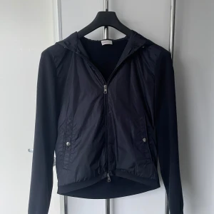 Moncler cardigan med huva - Snygg svart cardigan jacka från Industries med dragkedja och huva. Jackan har långa ärmar och två fickor med knappar framtill. Perfekt för en stilren look. Skicket är mycket bra. För fler frågor eller bilder kom privat!