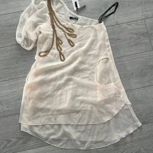 Champagnefärgad oneshoulder klänning från TFNC London - Elegant champagnefärgad oneshoulder klänning från TFNC London med en unik kedjedetalj i guld. Klänningen har en luftig passform och kort längd, perfekt för en stilfull kväll. Den har en transparent look med ett lager av lätt tyg med fickor 