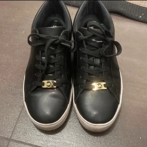 Svarta sneakers från Michael Kors - Snygga svarta sneakers från Michael Kors med gulddetaljer och snörning. Skorna har en rund tå och en vit sula som ger en stilren kontrast. Perfekta för en trendig look.