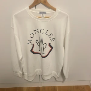 Vit sweatshirt från Moncler - Säljer en stilren vit sweatshirt från Moncler med deras ikoniska logga tryckt på framsidan. Tröjan har långa ärmar och en rund halsringning. Perfekt för en avslappnad och trendig look.
