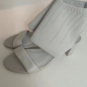 Grå sandaler med stickad detalj BE:GOLD - Unika grå sandaler från BE:GOLD med bred rem i skinn och stickad, ribbad sockliknande överdel. Skorna har öppen tå och låg klack, vilket ger en modern och cool vibe. Perfekta för dig som vill sticka ut med en trendig och bekväm look.