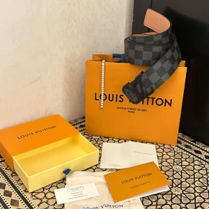 Fin rutig bälte  - Snyggt i klassiskt Damier Graphite-mönster. Bältet är svart och grått med logga på spännet och är tillverkat i exklusivt skinn. Perfekt accessoar för att lyfta din outfit med en lyxig touch.