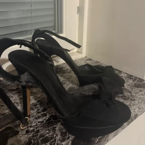 Svarta klackade sandaler med rosett - Svarta sandaler från MNG med hög klack och plattform, finns slitningar men det är enbart synligt på insidan av skon så de är inget man ser när man har på sig de🌸
