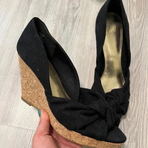 Svarta kilklackar från H&M  - Snygga svarta pumps från H&M med kilklack i kork och dekorativ rosett framtill. Skorna har öppen tå och är klädda i ett mjukt tygmaterial. Perfekta för dig som vill ha både höjd och komfort i en stilren look.