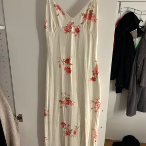 Vit långklänning med blommigt mönster - Superfin långklänning i vit ton med rosa och röda blommor. Klänningen har smala axelband, v-ringning och figurnära passform. Perfekt för sommarens alla tillfällen. Materialet känns mjukt och svalt mot huden. Endast använd 1 gång på en vin sommarkväll 🥰💗💗