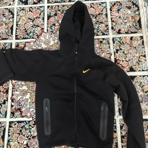 Nocta x Nike - Svart hoodiejacka från Nike med gul logga på bröstet och detaljer i gult på ryggen. Jackan har hel dragkedja framtill, två fickor med dragkedja och en stor huva. Snygg och sportig look, perfekt för chill eller träning.