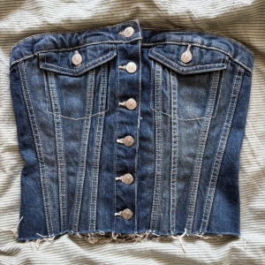 Jeanskorsett från Zara med knappar - Säljer en snygg jeanskorsett från Zara i mellanblå denim med silverfärgade knappar framtill, två fejkfickor och rå, fransig nederkant. Baksidan har smock för extra stretch och passform. Perfekt att styla med kjol eller byxor för en cool look.