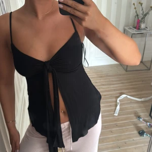 Svart linnetopp med knyt fram - Svart linnetopp med smala axelband och snygg knytning framtill som ger en öppen detalj. Tunt och mjukt material som känns skönt mot huden. Perfekt för varma dagar och enkel att styla med både jeans och kjol.