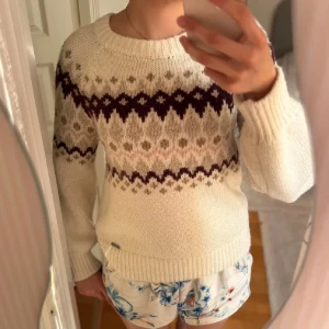 Vit stickad tröja med mönster - Supermysig vit stickad tröja med klassiskt mönster i mörklila och beige. Tröjan har rund halsringning, långa ärmar och ribbade muddar. Perfekt för kalla dagar när du vill vara både varm och snygg.