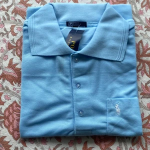 Ljusblå pikétröja från Ralph Lauren - Snygg ljusblå pikétröja från Ralph Lauren med klassisk krage och tre knappar framtill. Tröjan har det ikoniska broderade polospelar-loggan i vitt på bröstet. Perfekt för en fräsch och stilren look.