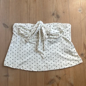 Vit prickig tubtopp med knyt - Supersöt vit tubtopp med svarta prickar och knyt framtill. Toppen är bandeau-modell och har en lätt rynkad detalj vid bysten. Aldrig använd för tyckte inte den passade på mig, däremot tvättad en gång❣️