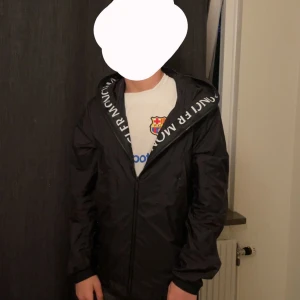 Moncler jacka/ windbreaker  - Säljer en stilren och exklusiv vindjacka från Moncler i svart, med dragkedja och en rejäl huva där Moncler-loggan är tryckt längs kanten – riktigt snygg detalj! Jackan är i toppskick och knappt använd. Perfekt för både höst, vår och svala sommarkvällar. Unisex-modell som passar alla. Storlek 2=M  🌟 Nästan ny 🌟 Ikonisk design 🌟 Passar till allt  Äkthetsbevis kan självklart visas vid förfrågan. Priset kan diskuteras vid snabb affär 