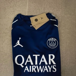 PSG x Jordan blå matchtröja - Snygg blå PSG fotbollströja från Jordan med vita detaljer och vingmönster på axlarna. Tröjan har klubbmärke på bröstet, Jordan-logga och Qatar Airways som sponsortryck. Tillverkad i lätt och ventilerande material, perfekt för fotbollsplanen. Pris kan diskuteras vid snabb affär 