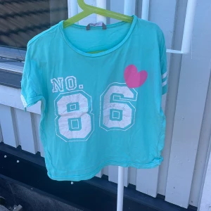 Turkos t-shirt med tryck från Lindex - Turkos t-shirt från Lindex med vitt tryck 'NO. 86' och en rosa hjärta på bröstet. T-shirten har korta ärmar med vita ränder och en avslappnad passform. Perfekt för en sportig och lekfull stil.