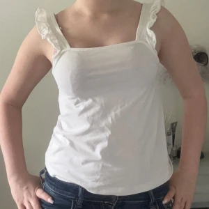 Vit linnetopp med volangaxelband - Supersöt vit linnetopp med raka linjer och breda axelband dekorerade med volanger. Toppen har en enkel och fräsch look, perfekt till jeans eller kjol. Tillverkad i mjuk bomull som känns skön mot huden. Axelbandsmodellen ger en feminin touch och passar perfekt för varma dagar.