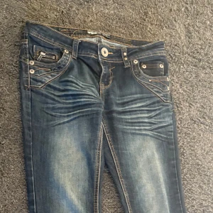 Blå jeans med detaljer och nitar - Säljer ett par blå jeans med slitningar och coola detaljer som nitar, dragkedja vid fickan och broderade bakfickor. Jeansen har kontrastsömmar och ett bälte i midjan. Perfekta för dig som gillar en unik och edgy stil. Pris kan diskuteras vid snabb affär 