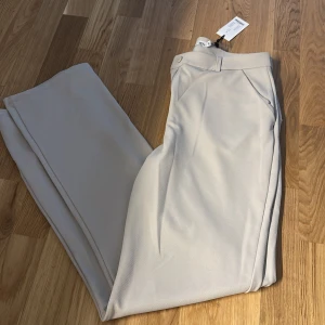 Beiga kostymbyxor med pressveck - Snygga beiga kostymbyxor från Vero Moda med klassiskt snitt och pressveck. Byxorna har hög midja, raka ben och stängs med knapp och dragkedja. Perfekta för en clean och stilren look.