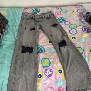 Grå slitna jeans från Gina Tricot bootcut  - Grå jeans från Gina Tricot i storlek 152 med coola slitningar och hål på benen. Baksidan har fransiga bakfickor och en vit stjärna på ena fickan. Jeansen har raka ben och klassisk femficksmodell. Perfekta för en avslappnad streetstil.