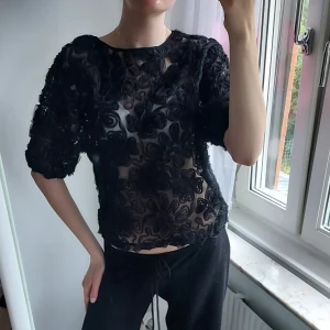 Svart blommig mesh blus - Säljer en svart transparent meshblus med stora 3D-blommor över hela tyget. Blusen har rund halsringning och korta puffärmar. Perfekt för dig som vill sticka ut med en unik och edgy look.
