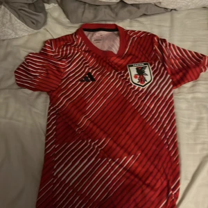 Japan Adidas röd fotbollströja S - Riktigt snygg Japan fotbollströja från Adidas i rött med vita diagonala ränder och svart logga. Tröjan har korta ärmar, rund hals och är i slim fit-modell. Materialet är lätt och andas, perfekt för match eller träning. JFA-emblem på bröstet.